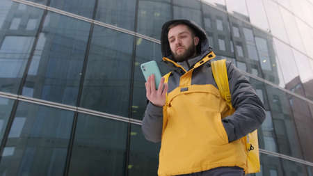 Unshaven courier with yellow backpack on his back in uniform walks down street in winter among tall...