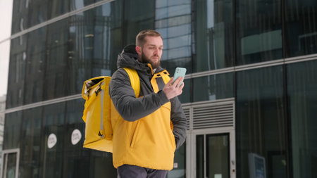 Unshaven courier with yellow backpack on his back in uniform walks down street in winter among tall...