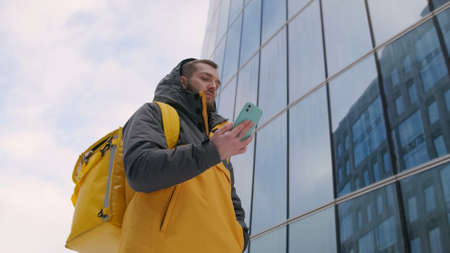 Unshaven courier with yellow backpack on his back in uniform walks down street in winter among tall...