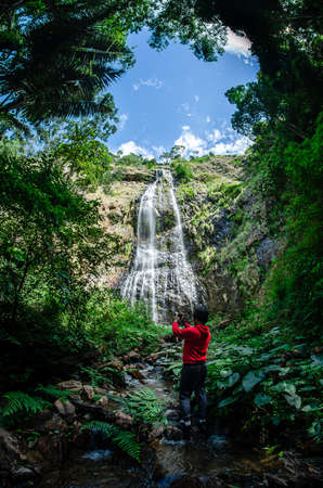asia man taking photos of waterfall with his cellphone. Male hiker photographing a water fall in forest.の写真素材