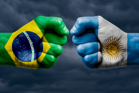 BRAZIL vs ARGENTINA confrontation, religious conflict. Men's fists with painted flags of BRAZIL and ARGENTINA. Dark clouds backgroundの写真素材