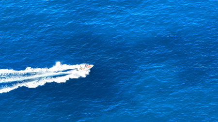 A small boat glides rapidly across the shimmering blue ocean, leaving a trail of white foam behind. The scene captures the beauty of the water on a sunny day.の写真素材