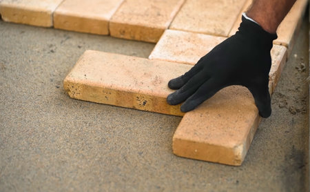 A worker in a black glove carefully places a paving stone onto a sandy surface. The individual is focused on aligning the stone as part of an outdoor construction project, showing attention to detail.の写真素材
