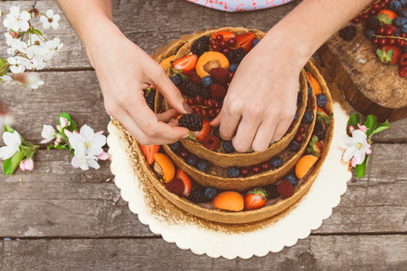 Woman hands of baker carefully decorate cheesecake with fruits and berries on the old planks, white apple and cherry flowersの写真素材
