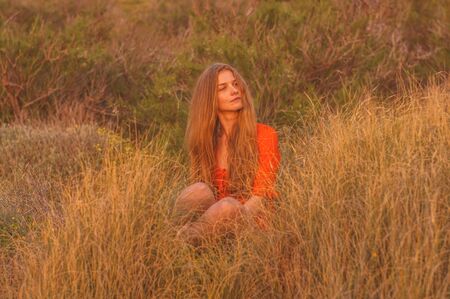 Young blonde girl in red dress sitting on grass in the eveningの写真素材