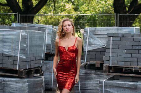 Beautiful blond girl in red short dress with sequins in park near construction site