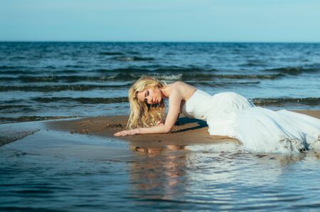 Portrait of young blond bride lies on sand among the water, looking to the cameraの写真素材