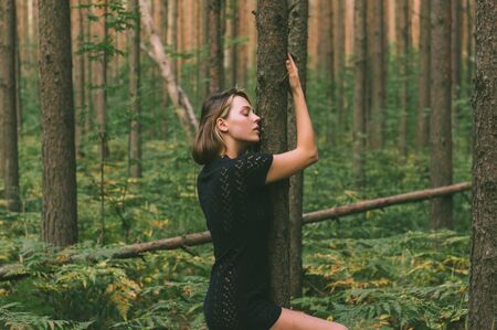 Portrait of young beautiful girl in black dress in forest that holds tree trunkの写真素材
