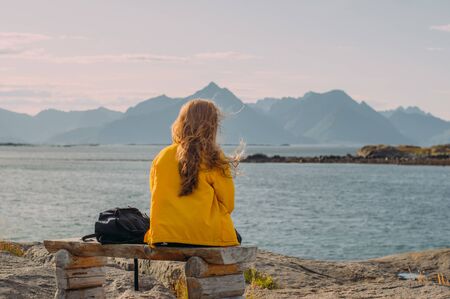 Girl in yellow jacket sitting on the bench and looking to mountains and and sea and fjord, hiking in hillsの写真素材