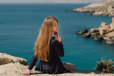 Beautiful smiling blond girl  with strawberry in hand sitting on the edge of mountain with view to the sea, side viewの写真素材