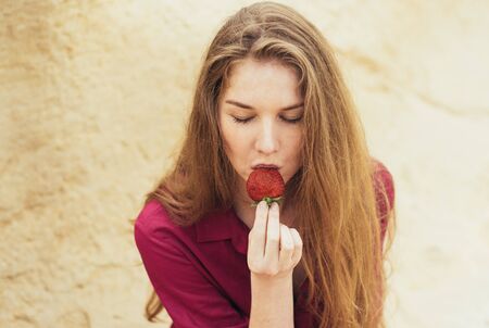 Beautiful blond smiling girl in purple dress eating fresh strawberry on natureの写真素材