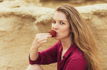 Beautiful blond smiling girl in purple dress eating fresh strawberry on natureの写真素材