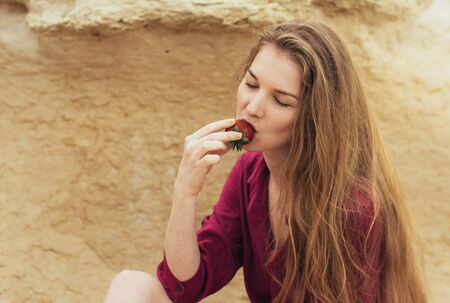 Beautiful blond girl in purple dress eating fresh strawberry on natureの写真素材