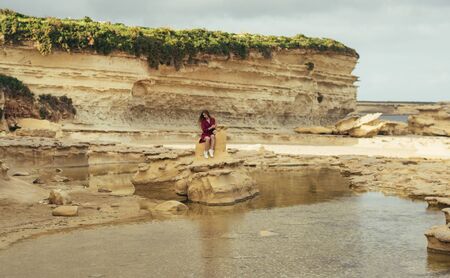 Beautiful blond smiling girl in purple dress sitting on stone on natureの写真素材