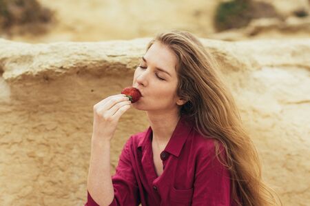Beautiful blond girl in purple dress eating fresh strawberry on natureの写真素材
