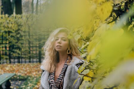 Young blond girl staying near trees in public park in fall and looking to camera