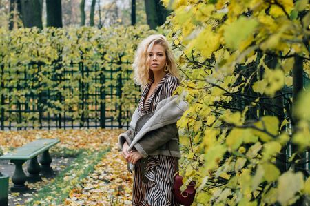Young blond girl staying near trees in public park in fall and looking to cameraの写真素材
