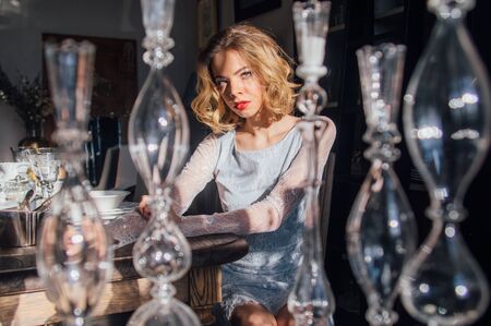 Beautiful blond girl with vintage make up and hairstyle sitting near table with sunny rays on face, glass candlesticks on foregroundの写真素材