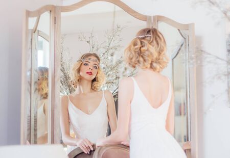 Beautiful blond girl with vintage make up and hairstyle staying near mirror with jewelry on head and looking to cameraの写真素材