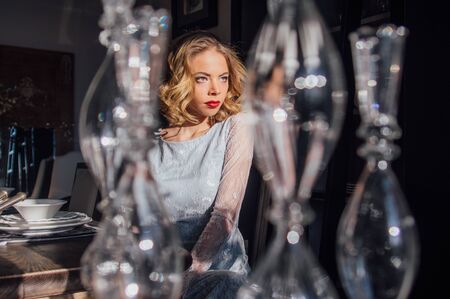 Beautiful blond girl with vintage make up and hairstyle sitting near table with sunny rays on face, glass candlesticks on foregroundの写真素材