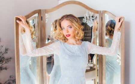 Beautiful blond girl with vintage make up and hairstyle staying near triple mirror and looking to...