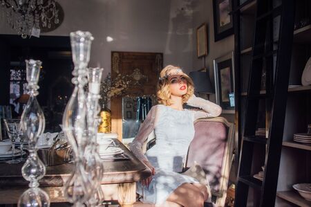 Beautiful blond girl with vintage make up and hairstyle sitting near table with sunny rays on face and jewelry decoration on head, glass candlesticks on foregroundの写真素材