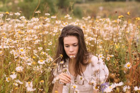 Young beautiful girl in white dress smelling flowers in chamomile field in summerの写真素材