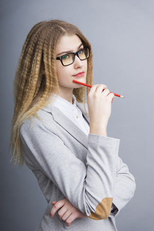 Young thoughtful business woman in an official suit and glasses holding pencil standing on the gray backgoundの写真素材