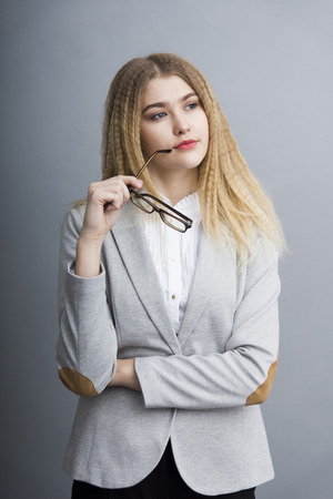 Young thoughtful business woman in an official suit holding glasses standing on the gray backgoundの写真素材