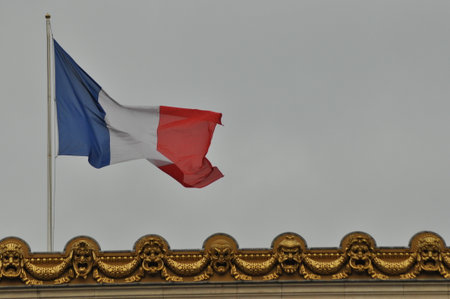 The French flag flutters atop an ornate building in Paris. The cloudy sky creates a dramatic backdrop, emphasizing the national pride displayed by the vibrant colors of the flag.の写真素材