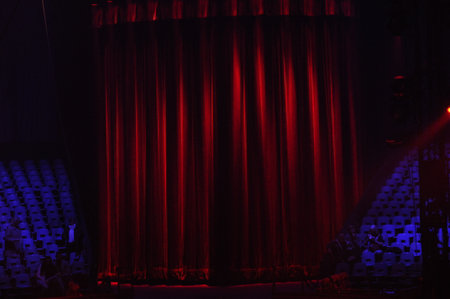 Red curtains hang on the stage in a dimly lit theater, creating a sense of anticipation. Empty seats await the audience, ready for an upcoming show.の写真素材