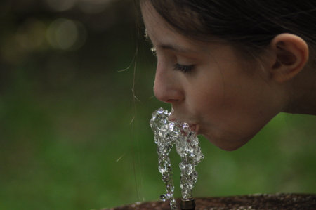 A young girl leans over a garden fountain, sipping clear water as it flows. Sunlight filters through...