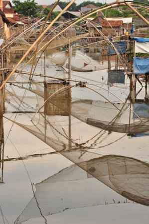 In a tranquil rural village, wooden structures and large fishing nets stretch over calm waters at dusk. The scene captures the peaceful rhythm of village life and traditional fishing techniques.の写真素材