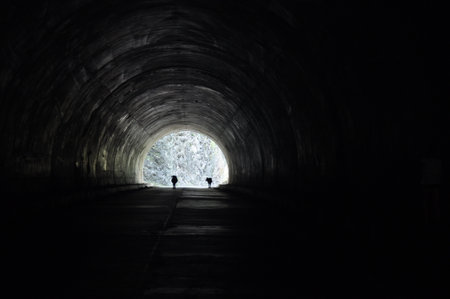 Two hikers venture through a dark tunnel, their silhouettes contrasting against a bright light at the end. The scene captures a moment of exploration and adventure.の写真素材