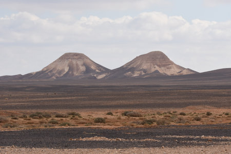 Twin mountains dominate the stark desert vista, showing their smooth slopes against a backdrop of clouds. The terrain is dry and rocky, highlighting the natural beauty of isolation.の写真素材