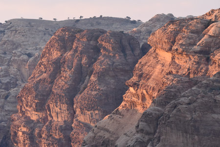 Towering rock cliffs are illuminated by the soft glow of the setting sun, highlighting the rugged terrain in a desert landscape. The scene captures the beauty of nature at dusk.の写真素材