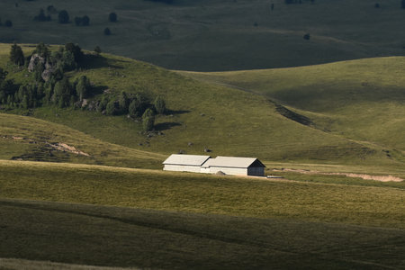 A spacious countryside features a rustic barn surrounded by rolling green hills. Sunlight casts...