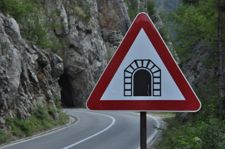 A caution sign warns drivers of an approaching tunnel as the scenic road curves through rocky hills covered in greenery during daylight hours.の写真素材