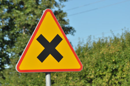 A triangular warning sign with a black cross stands tall beside a road. Lush green trees can be seen...