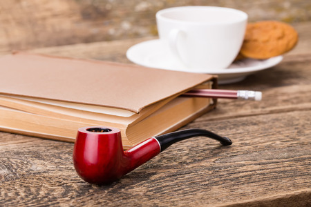Tobacco pipe for smoking, ivory tea cup with sweet cookie, and book on wooden paletteの写真素材