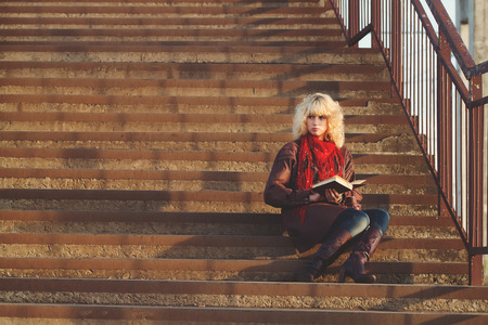 Young girl in leather coat with book seat on stairs bridge in sunshine, sunriseの写真素材