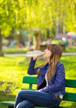 Beautiful Fashion Woman with handbag sitting and waiting on a bench in the park and drink waterの写真素材