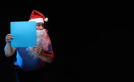 portrait actor male in cap and beard of Santa Claus with a sheet of paper for notes in the hands on a black backgroundの写真素材