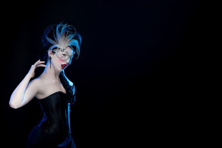 brunette woman with high hair, a mask with feathers and corset in old style on a black background