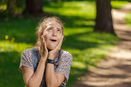 Emotional teenager girl with long hair in blue denim shorts walks in the park on a sunny day.