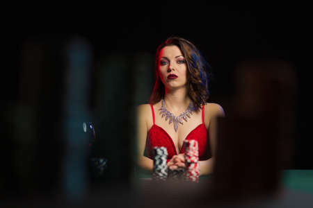 Emotional woman in an evening red dress playing cards with money on the table on a green cloth in a...