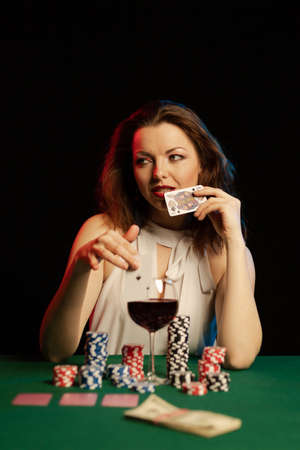 Emotional young lady in a white blouse drinking wine from a glass and playing cards on a table on green cloth in a casinoの写真素材
