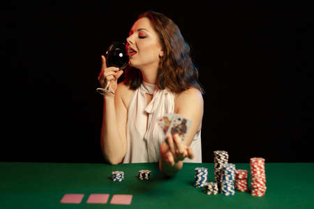 Emotional young lady in a white blouse drinking wine from a glass and playing cards on a table on...