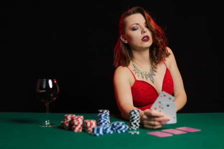 Emotional young girl in an evening red dress playing cards with money on the table on a green cloth in a casinoの写真素材