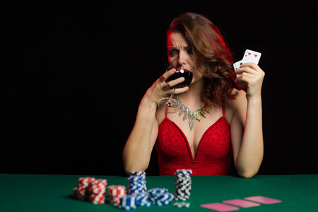 Emotional young woman in an evening red dress playing cards with money on the table on a green cloth...